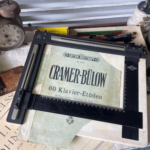May include: An antique still life featuring a vintage clock, sheet music, and a drafting tool. The sheet music is titled "Cramer-Bülow 60 Klavier-Etüden". The clock has a round face with black numbers. Pencils and other items are also visible.