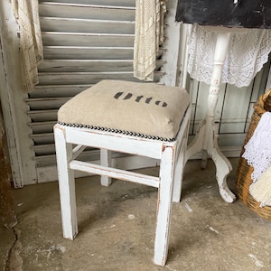 Può includere: Un piccolo sgabello in legno bianco con un cuscino rettangolare beige. Il cuscino è stampato con la parola "Otto" in lettere nere. I bordi del cuscino sono rifiniti con piccole borchie nere. Lo sgabello è in stile vintage.