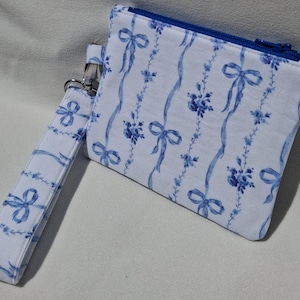 May include: A white fabric wristlet pouch featuring a repeating blue pattern of delicate floral sprigs and tied ribbons. The rectangular pouch includes a matching wrist strap attached by a silver metal clasp and a blue zipper closure at the top.