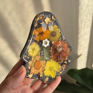 May include: A ghost-shaped resin ornament featuring a dark, translucent base embedded with various pressed flowers in shades of yellow, orange, and brown, accented with small gold leaf flakes throughout the clear, glossy, and smooth surface.