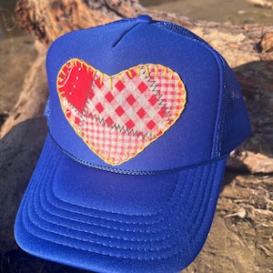 May include: A royal blue trucker hat featuring a large, heart-shaped patch on the front. The heart is constructed from red and white gingham fabric pieces stitched together with yellow thread, creating a patchwork design with a visible zigzag seam.