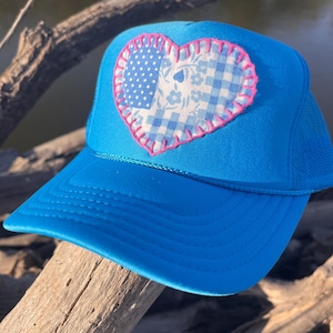 May include: A bright blue trucker hat featuring a heart-shaped patch on the front panel. The patch is constructed from a patchwork of light blue and white fabric patterns, including polka dots, floral prints, and gingham, outlined with pink stitching.