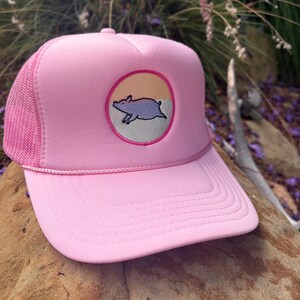 Könnte beinhalten: Eine leuchtend rosa Trucker-Cap mit Schaumstoff-Vorderseite und Netzrückseite. Die Kappe hat einen runden Aufnäher auf der Vorderseite, der ein hellviolettes Schwein vor einem geteilten pfirsichfarbenen und grauen Hintergrund zeigt, mit Kordel.
