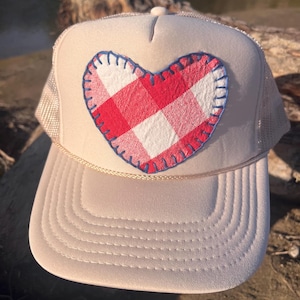 Op de afbeelding: Een beige truckerpet met een mesh achterkant en een gestructureerd voorpaneel. In het midden aan de voorzijde bevindt zich een stoffen hartvormige patch met een rood-wit ruitpatroon, omlijnd met blauwe stiksels en een dun koord.