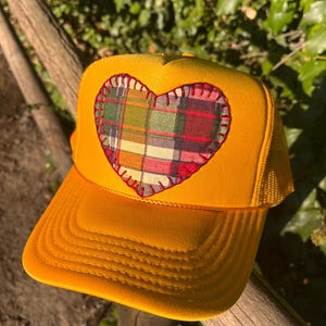 Op de afbeelding: Een felgele truckerpet met een hartvormige patch op het voorpaneel. De patch is gemaakt van een veelkleurige ruitjesstof in tinten rood, groen en geel, vastgezet met zichtbare rode festonsteken langs de randen van het hart.