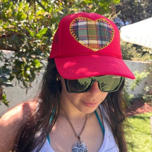 May include: A bright red trucker hat featuring a large heart-shaped patch on the front. The heart is made of a multi-colored plaid fabric in shades of green, blue, and yellow, and is outlined with thick, decorative yellow stitching.