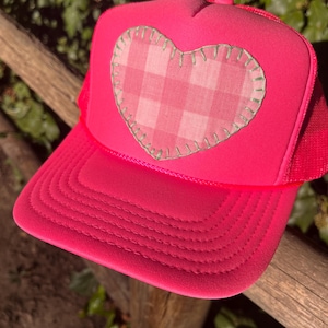 Op de afbeelding: Een fel neonroze truckerpet met een grote, centraal geplaatste hartvormige patch. Het hart is gemaakt van lichtroze en witte gingham-stof, omlijnd met zichtbare saliegroene stiksels. De pet heeft een gestructureerd voorpaneel en mesh.