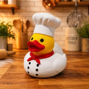 Peut inclure: Un canard en caoutchouc jaune portant une toque de chef blanche et une veste de cuisine blanche avec un foulard rouge. Le canard a un bec rouge vif, des yeux noirs et six petits boutons noirs sur le devant de la veste, formant une figurine.