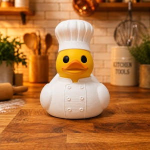 Peut inclure: Un canard en caoutchouc jaune portant une toque de chef blanche et une veste de cuisine blanche à double boutonnage avec de petits boutons circulaires. Le canard a un bec orange vif et des yeux noirs, présenté de face avec une texture mate.