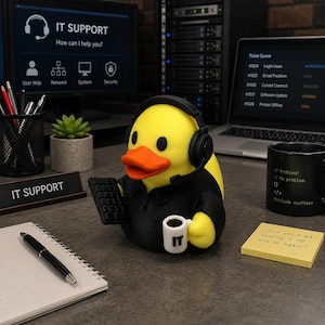 Könnte beinhalten: Eine gelbe Quietscheente-Figur in einem schwarzen Hemd mit Kragen, die einen schwarzen Kopfhörer mit Mikrofon trägt. Die Ente hält eine kleine schwarze Tastatur in einem Flügel und eine weiße Tasse mit der Aufschrift IT im anderen Flügel.