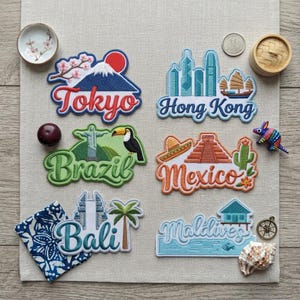 May include: Six embroidered patches featuring travel themes: Tokyo with Mount Fuji, Hong Kong skyline, Brazil with Christ the Redeemer and a toucan, Mexico with a pyramid and cactus, Bali with a temple and palm tree, and the Maldives with a water villa.