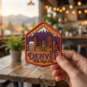 May include: A hexagonal embroidered patch featuring a purple and orange Denver skyline with a white mountain peak, a beer mug, and a hiking boot. The text reads DENVER Mile High City in purple and black lettering with a bright orange border.