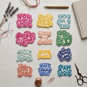 Puede incluir: Una colección de doce parches bordados con frases motivacionales que incluyen You Are Brave, You Are Enough, You Can Do This, One Day At A Time, You Are Awesome, You Are Strong, You Matter, Be Kind To Yourself, Good Vibes, You Are Loved, Make Today Great e I Am Brave.
