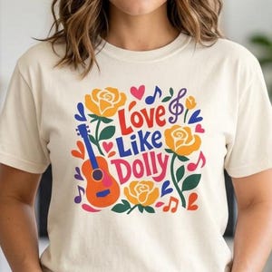 Könnte beinhalten: Ein cremefarbenes Kurzarm-T-Shirt mit einem bunten Grafikdruck und dem Text "Love Like Dolly". Das Design zeigt eine rote und orangefarbene Akustikgitarre, gelbe Rosen, grüne Blätter, blaue Musiknoten sowie kleine rote Herzen auf der Vorderseite.