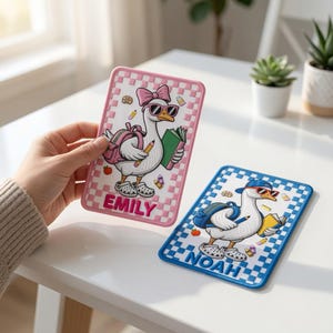 May include: Two rectangular embroidered patches featuring cartoon geese. The pink-bordered patch shows a goose with a bow, sunglasses, and backpack, holding a book and pencil, with the name EMILY. The blue-bordered patch shows a goose with a cap, sunglasses, and backpack, holding a book and pencil, with the name NOAH.