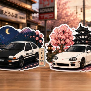May include: Two die-cut stickers featuring Japanese sports cars. The left sticker shows a white and black AE86 Trueno with Mount Fuji and a crescent moon. The right sticker shows a white Supra MK4 with a castle and cherry blossoms.