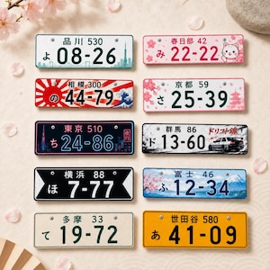 May include: Ten rectangular metal license plates featuring various Japanese designs, including cherry blossoms, Mount Fuji, a cat, a sports car, and city skylines. Each plate displays unique Japanese characters and numerical sequences in diverse colors.
