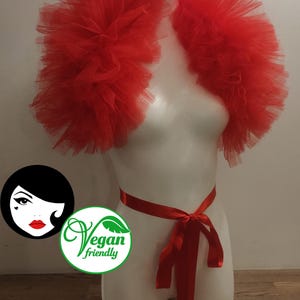 May include: A bright red tulle shrug featuring voluminous, layered ruffles that drape over the shoulders. The garment is secured at the waist with a matching red satin ribbon tied in a bow. A circular logo with the text Vegan friendly is visible.