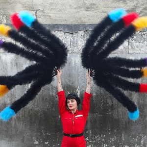 May include: A person wearing a red jumpsuit and a black beret holds two large black feather fans with colourful tips. The fans are spread wide, creating a dramatic effect.