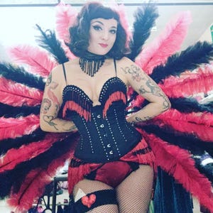 May include: A woman wearing a black corset with red fringe and a red and black feather boa. She is wearing a black choker necklace and has red lipstick on.