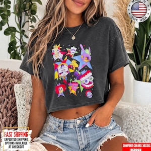 May include: A charcoal gray short-sleeve t-shirt featuring a vibrant graphic print of various cartoon characters, including a jester, a rabbit, and a toothy creature, set against colorful star shapes. The shirt has a crew neckline and a relaxed fit.
