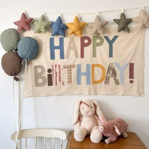Può includere: Uno striscione in tessuto con il testo HAPPY BIRTHDAY in lettere a motivi multicolori, appeso con una ghirlanda di stelle di peluche. Tre palloncini di peluche verdi, blu e marroni pendono a sinistra. Un coniglio e un maiale di peluche sono seduti.