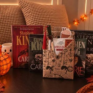 Puede incluir: Una colección de libros de Stephen King incluyendo Carrie, una funda de tela para libros con estampado de llaves antiguas, un marcapáginas con borla roja, un sobre de mezcla de sidra de manzana y una luz con forma de calabaza, con decoración otoñal.
