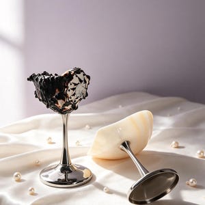 May include: Two stemmed dessert cups with metallic silver bases. One cup features a dark, swirled chocolate and cream interior, while the other is a smooth, pale cream-colored bowl. Both have polished silver stems and circular bases.