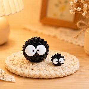 May include: Two spherical black soot sprite figurines with textured, spiky surfaces and large, round white eyes with black pupils. The larger figurine is positioned to the left, while the smaller, identical figurine sits to its right.