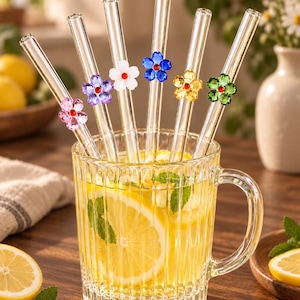 May include: Six clear glass drinking straws, each featuring a small, colourful five-petalled flower accent with a red centre. The straws are arranged inside a ribbed glass mug filled with a yellow citrus beverage containing lemon slices and mint leaves.