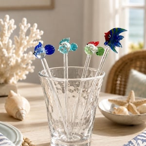 May include: A set of four clear glass drink stirrers, each topped with a small, colorful glass fish figurine. The fish are colored in shades of blue, light blue, green, and a multi-colored combination of red, blue, and green, all held in a glass.