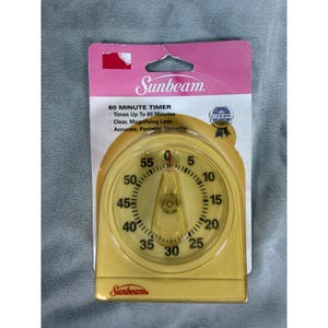 May include: A yellow Sunbeam 60 minute kitchen timer in its original packaging. The timer features a circular dial with black numbers from 0 to 55, a clear magnifying lens over the center, and the Sunbeam logo printed at the top and bottom.
