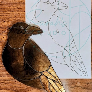 Puede incluir: Una figura de pájaro de vitral en tonos marrón oscuro y ámbar con líneas de plomo visibles, colocada junto a una plantilla de papel blanco con secciones numeradas y el texto Sarah's STUDIO en letra azul claro en el centro.