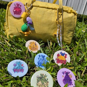 May include: A yellow handbag adorned with two keychains featuring charms like a lamp and beads, surrounded by seven circular buttons. Each button displays a colorful dress illustration with text including Fierce, Bold, Dream, Sweet, Fearless, Driven, and Wild.
