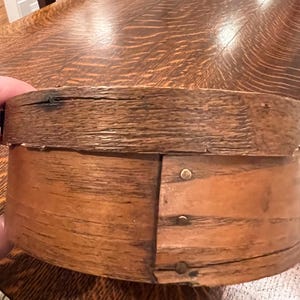 May include: A round wooden box with a textured, dark brown grain finish. The lid features a slightly overlapping edge, and the side of the base is secured with three small, circular metal fasteners arranged in a vertical line along the seam.