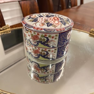 May include: A round, two-tiered porcelain container with a lid, featuring a detailed floral pattern in shades of red, blue, green, and white. The surface is decorated with intricate botanical motifs and geometric borders, showcasing a glossy finish.
