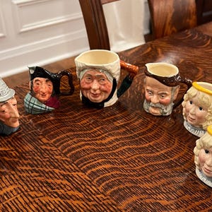 May include: A collection of seven ceramic character jugs and mugs featuring detailed hand-painted faces of various figures, including individuals in hats, a person with a white bonnet, a bearded man, and two blonde figures holding small harps.