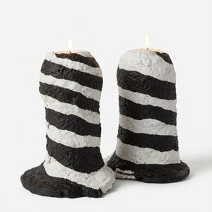 May include: Two pillar candles with a textured, uneven surface featuring alternating diagonal black and white stripes. Each candle has a wide, flared base and a lit wick at the top, creating a distinct, hand-molded appearance with visible wax depth.