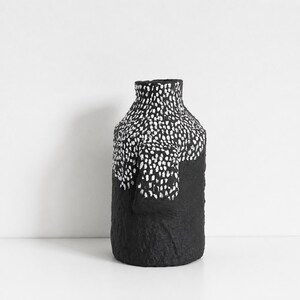May include: A cylindrical vase with a textured black surface. The upper portion features a dense pattern of small, white, hand-painted vertical dashes that transition into a solid black base. The vessel has a narrow neck and a flat rim.