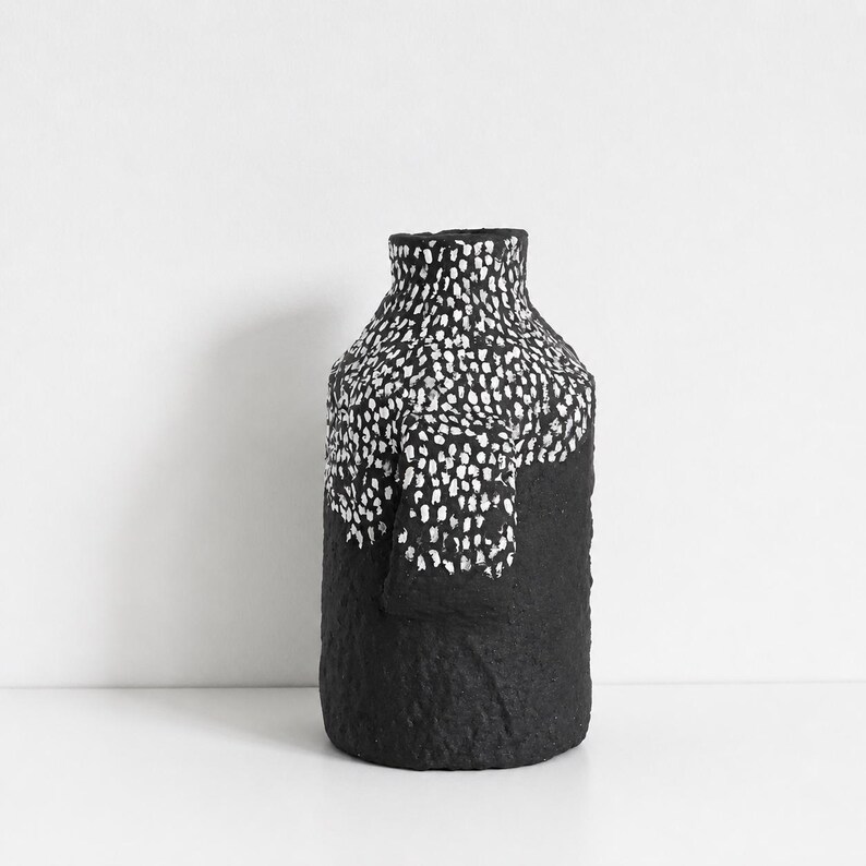 May include: A cylindrical vase with a textured black surface. The upper portion features a dense pattern of small, white, hand-painted vertical dashes that transition into a solid black base. The vessel has a narrow neck and a flat rim.