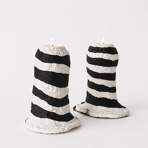 May include: Two cylindrical candles with a textured, uneven surface featuring horizontal black and white stripes. Each candle has a lit wick at the top, and the base of each candle flares outward with a rough, organic edge.