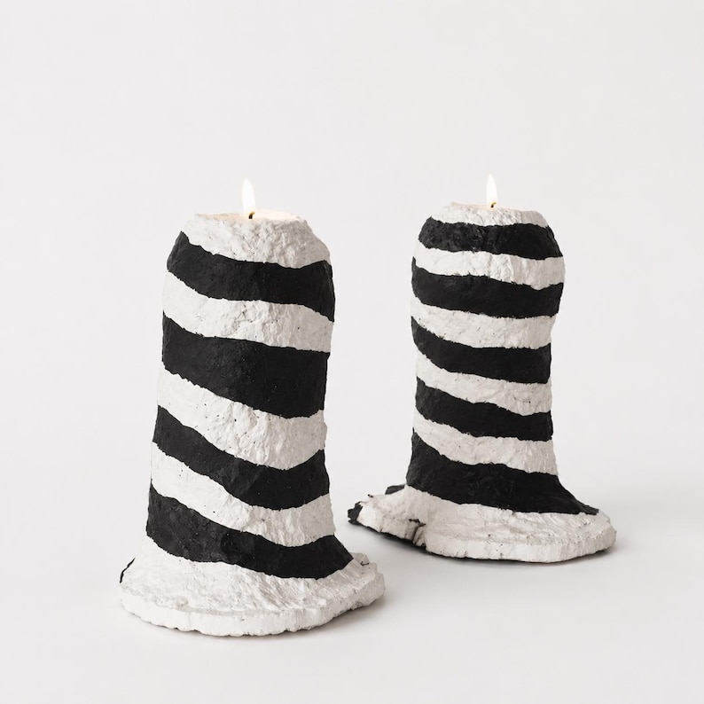May include: Two cylindrical candles with a textured, uneven surface featuring horizontal black and white stripes. Each candle has a lit wick at the top, and the base of each candle flares outward with a rough, organic edge.