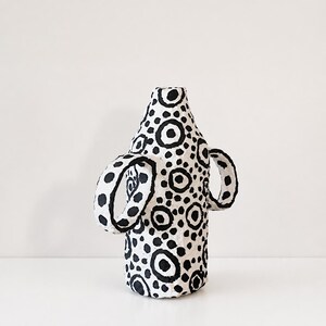 May include: A white vase with a textured surface featuring a hand-painted black pattern of various sized circles and dots. The vessel has a cylindrical body, a narrow neck, and two prominent, curved handles extending from the sides of the main body.
