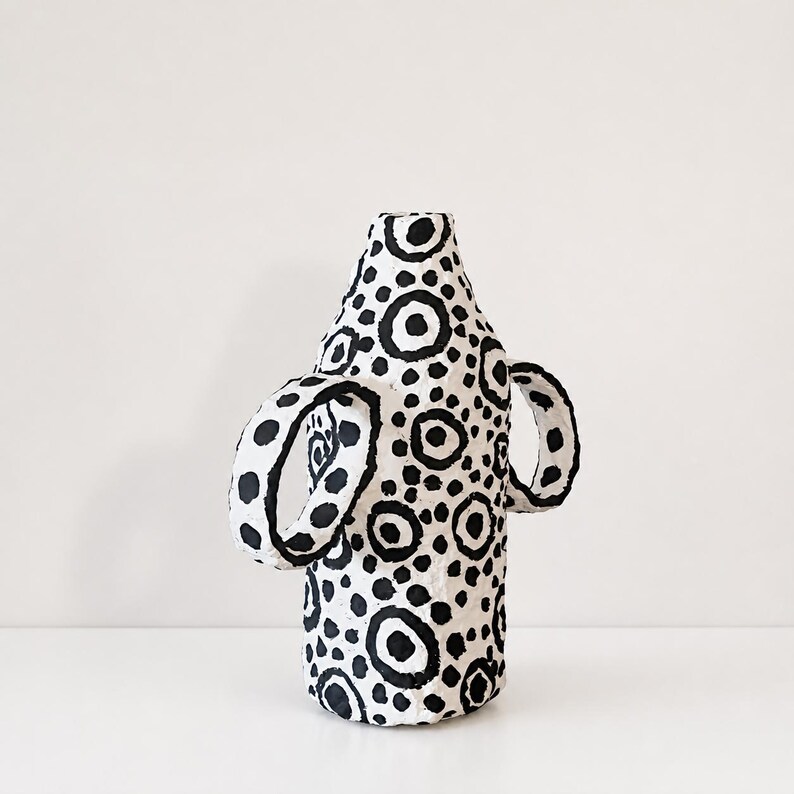 May include: A white vase with a textured surface featuring a hand-painted black pattern of various sized circles and dots. The vessel has a cylindrical body, a narrow neck, and two prominent, curved handles extending from the sides of the main body.