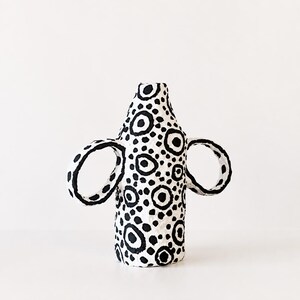 May include: A white ceramic vase with a textured surface featuring a hand-painted black pattern of various sized circles and dots. The vessel has a cylindrical body with a narrow neck and two large, circular handles extending from the sides.