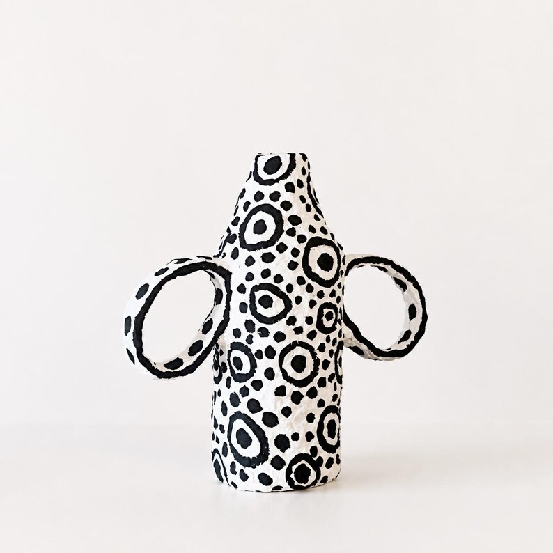 May include: A white ceramic vase with a textured surface featuring a hand-painted black pattern of various sized circles and dots. The vessel has a cylindrical body with a narrow neck and two large, circular handles extending from the sides.
