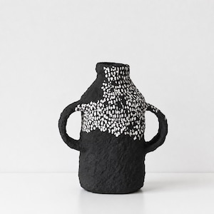 May include: A black vase with two side handles and a textured surface. The upper portion of the vase features a dense pattern of small white dots that contrast against the dark base, creating a distinct visual separation across the body.