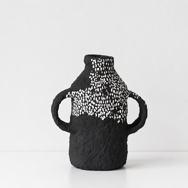 May include: A black vase with two side handles and a textured surface. The upper portion of the vase features a dense pattern of small white dots that contrast against the dark base, creating a distinct visual separation across the body.