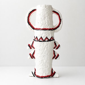 May include: A white textured vase with a rough, paper-like finish. The vessel features two large arched handles at the top and two pairs of smaller, pointed protrusions along the sides. It is decorated with hand-painted red and black lines.