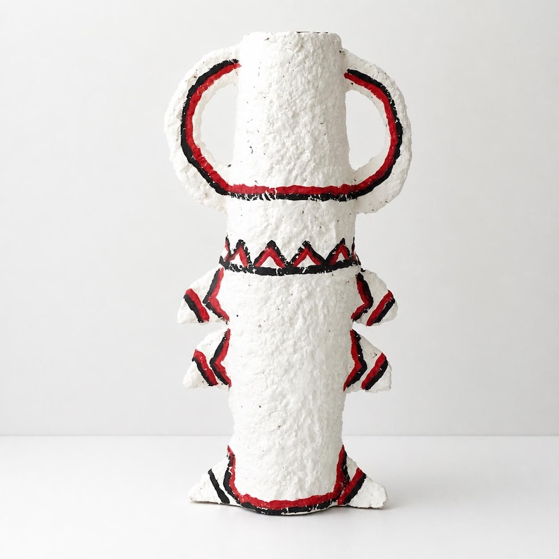 May include: A white textured vase with a rough, paper-like finish. The vessel features two large arched handles at the top and two pairs of smaller, pointed protrusions along the sides. It is decorated with hand-painted red and black lines.